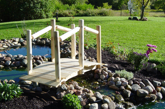 Wooden Decorative Garden Bridge over a small pond, surrounded by rocks, greenery, and flowers in a backyard.